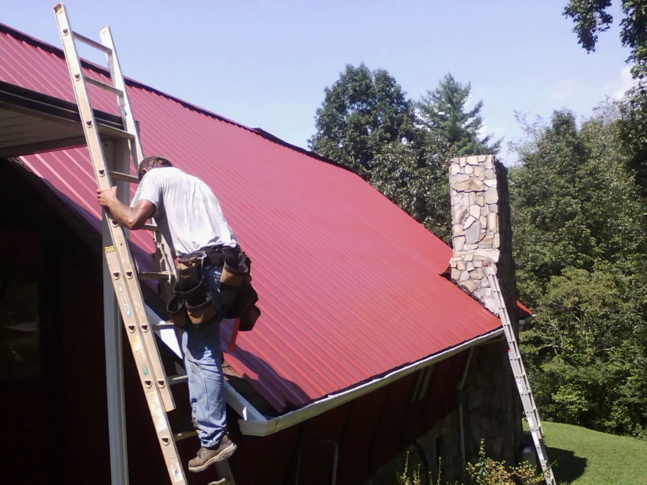 Roofer on a ladder beside a red metal roof and stone chimney — Soffit & Fascia Repair by Roofing Brothers in Star City